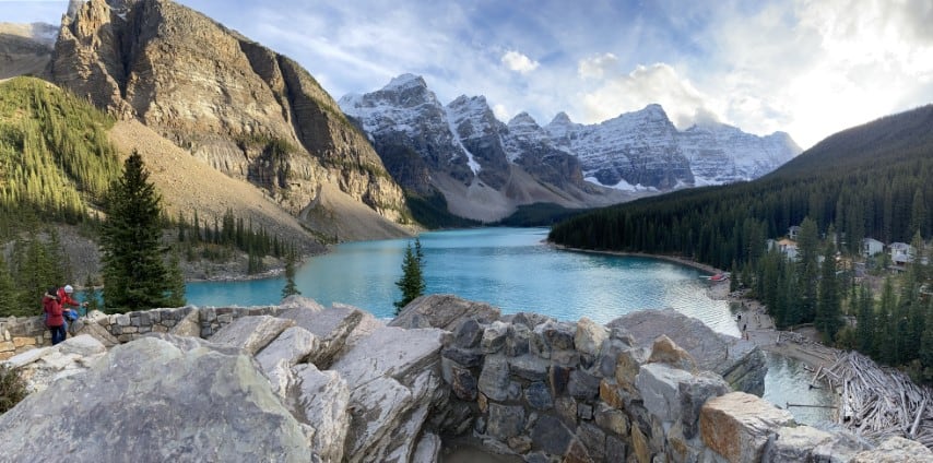 A photo of Lake Moraine just before fall. The sun is shining on the clouds and the closes peak is being lit up over the green glacial water of the lake.